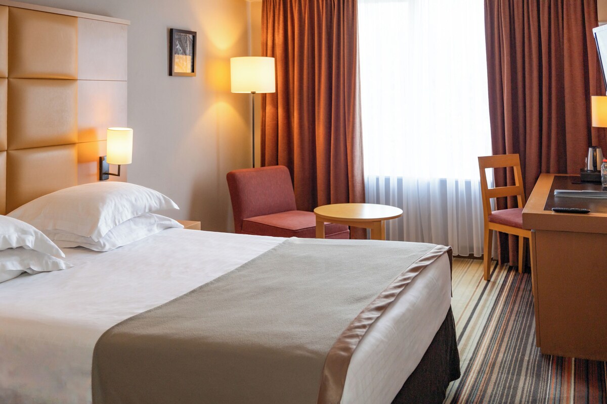 A hotel room with a bed, two chairs, a small table, a desk, and a lamp.