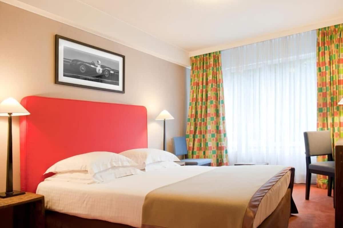 A hotel room with a bed, two bedside lamps, a chair, a desk, a framed picture on the wall, and a window with curtains.