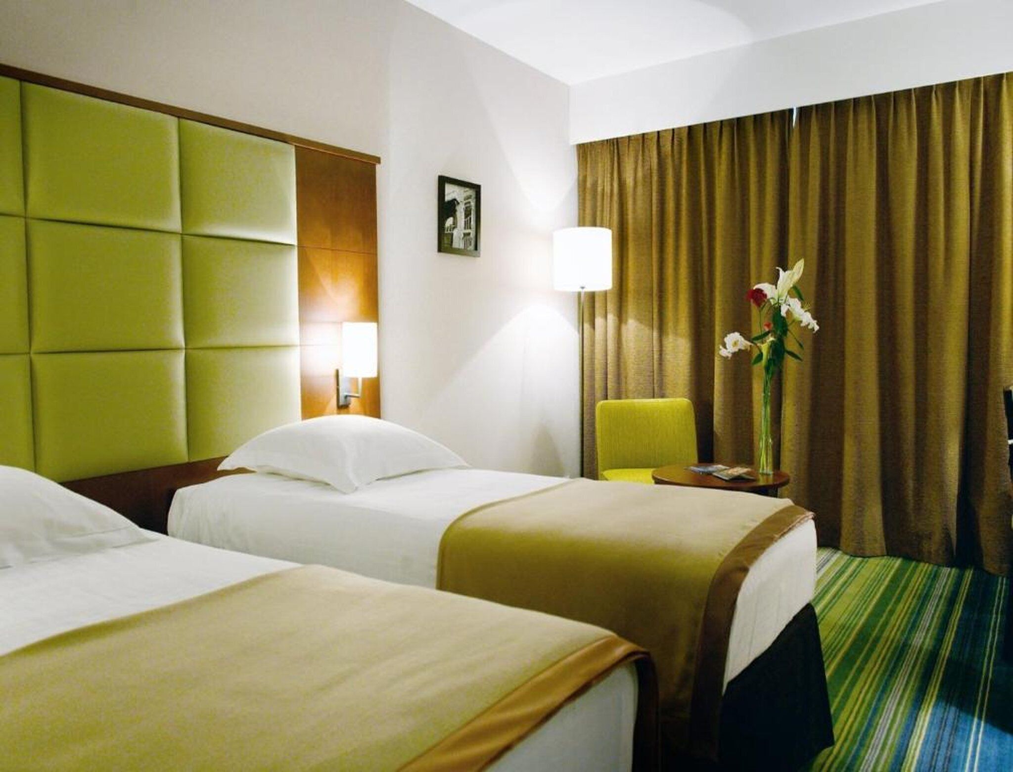 A hotel room with two beds, a green headboard, a lamp, a chair, a vase with flowers, and a window with curtains.
