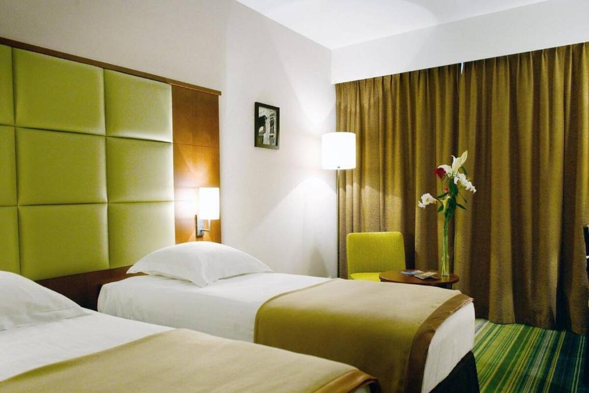 A hotel room with two beds, a green headboard, a lamp, a chair, a vase with flowers, and a window with curtains.