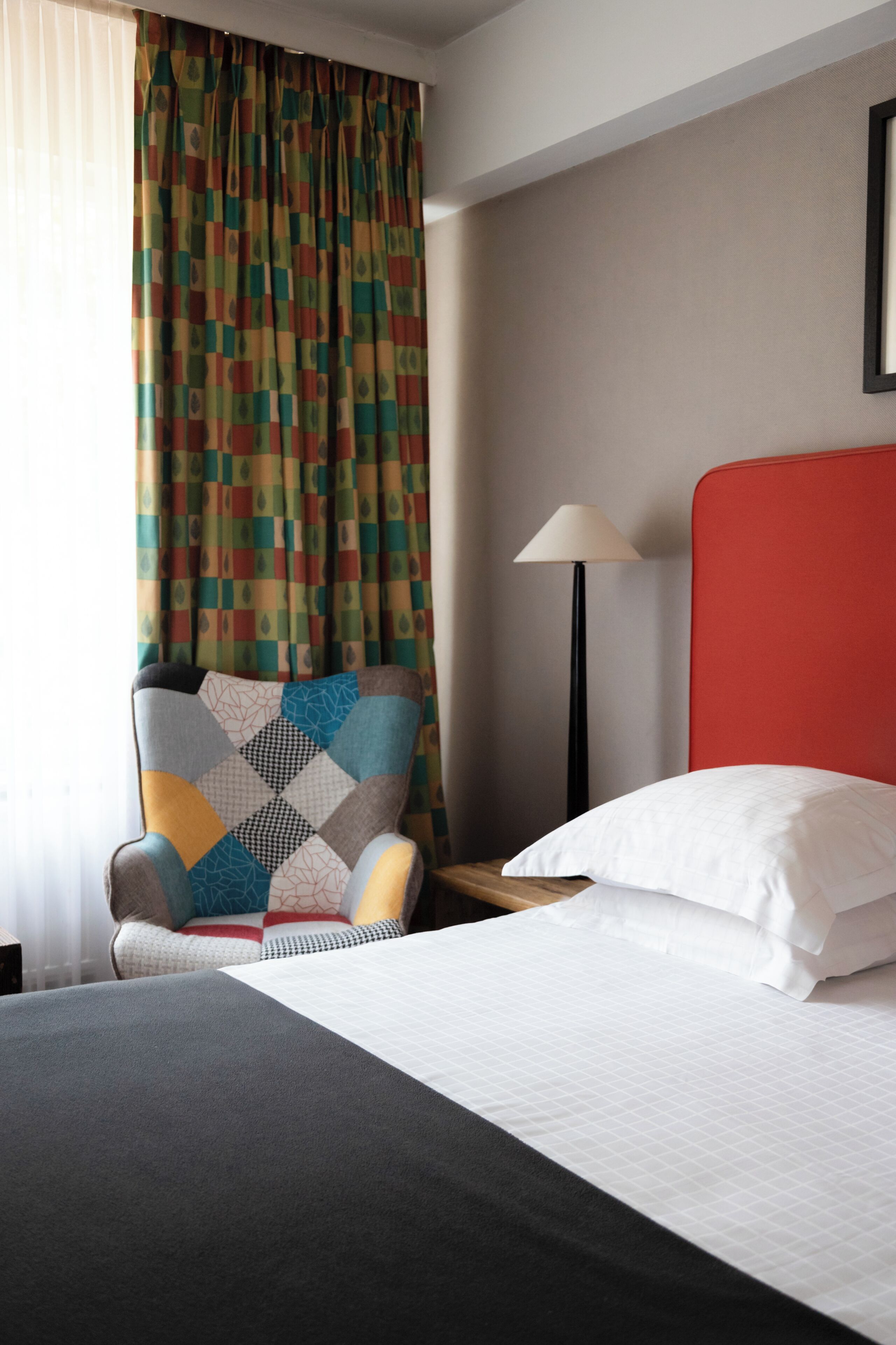 Habitación de hotel con una cama, una silla con cojín estampado, una lámpara y una ventana con cortinas.