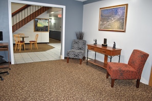 A room with a patterned carpet, a wooden desk, a chair, a patterned armchair, and a framed picture on the wall.