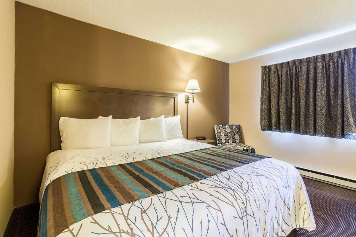 Een hotelkamer met een groot bed, een hoofdbord, een bedlampje, een gestreept fauteuil en een raam met gordijnen.