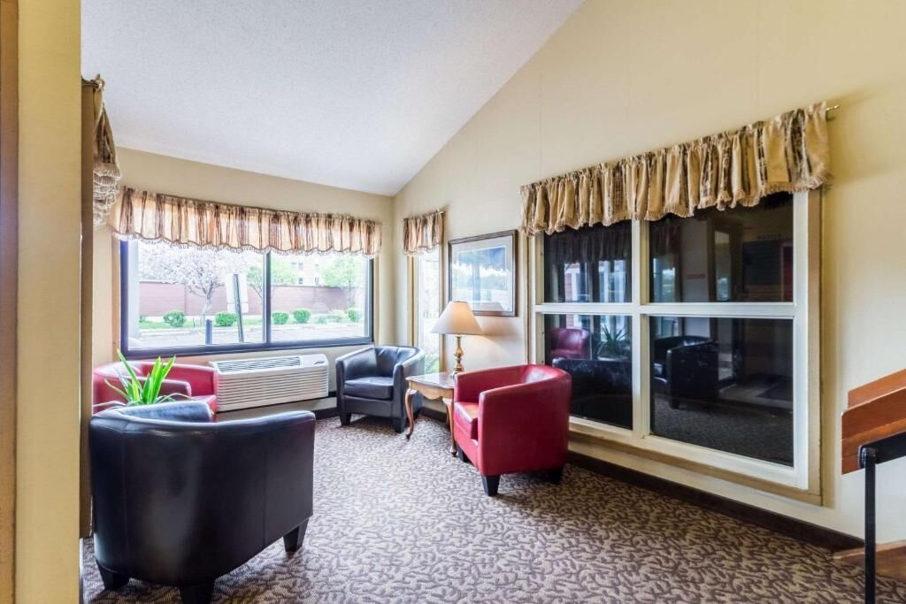 A hotel room with a large window, a fireplace, a red armchair, a black armchair, and a patterned carpet.