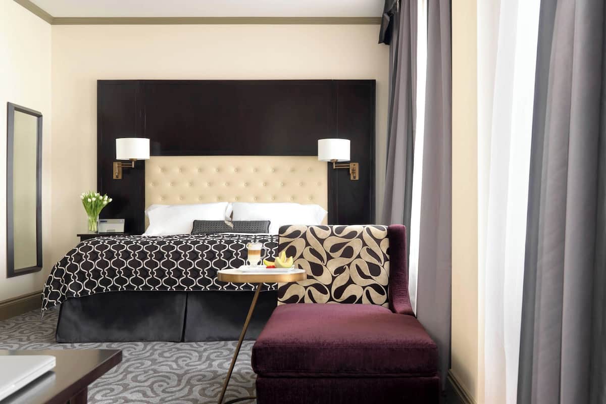 Et hotelværelse med en stor seng, to sengeborde med lamper, et lille bord med en drink, en vase med blomster og udsigt udendørs gennem gardiner.