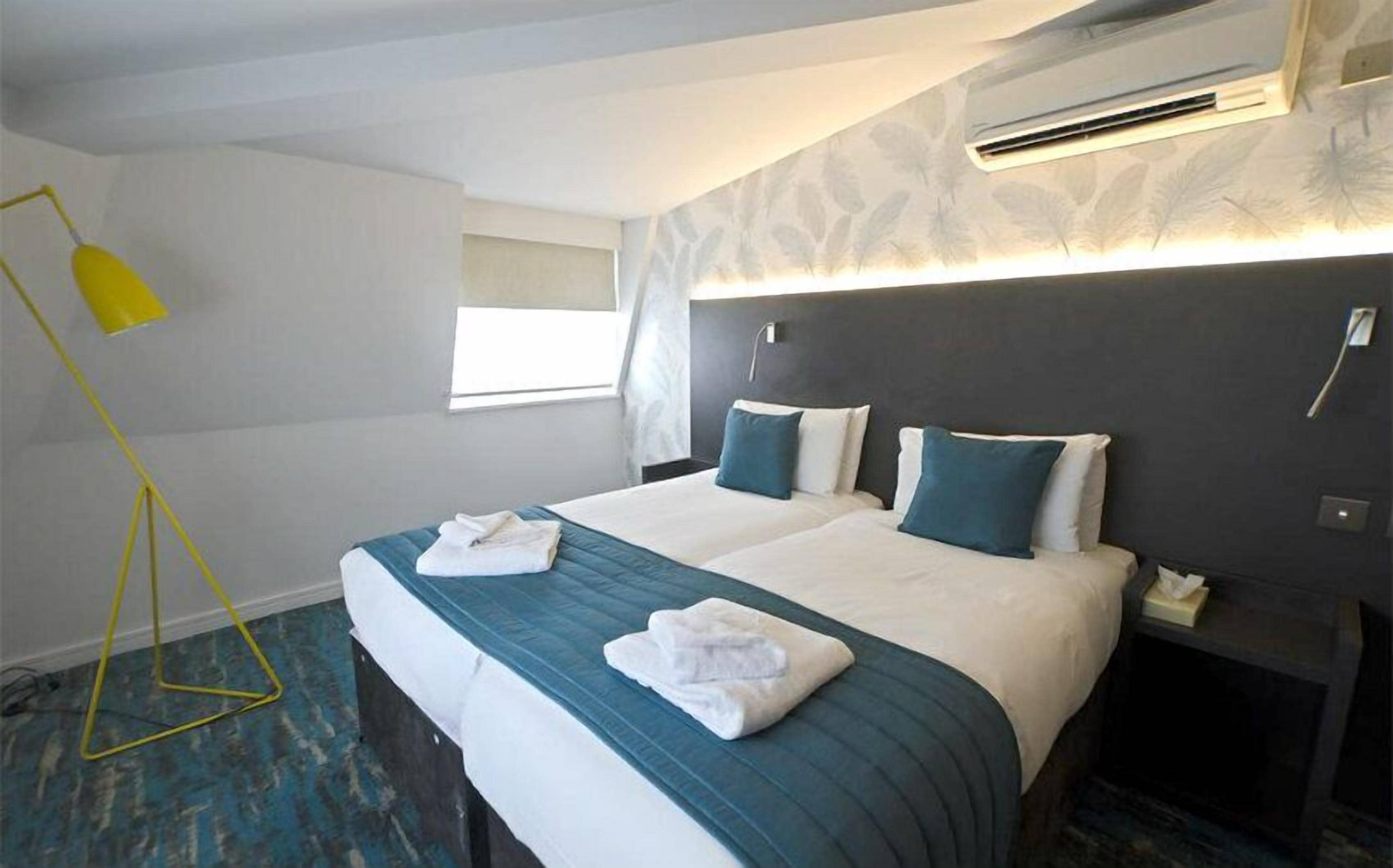 A hotel room with a bed, two pillows, a bedside table, an air conditioning unit, a window, and a floor lamp.