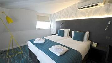 A hotel room with a bed, two pillows, a bedside table, an air conditioning unit, a window, and a floor lamp.