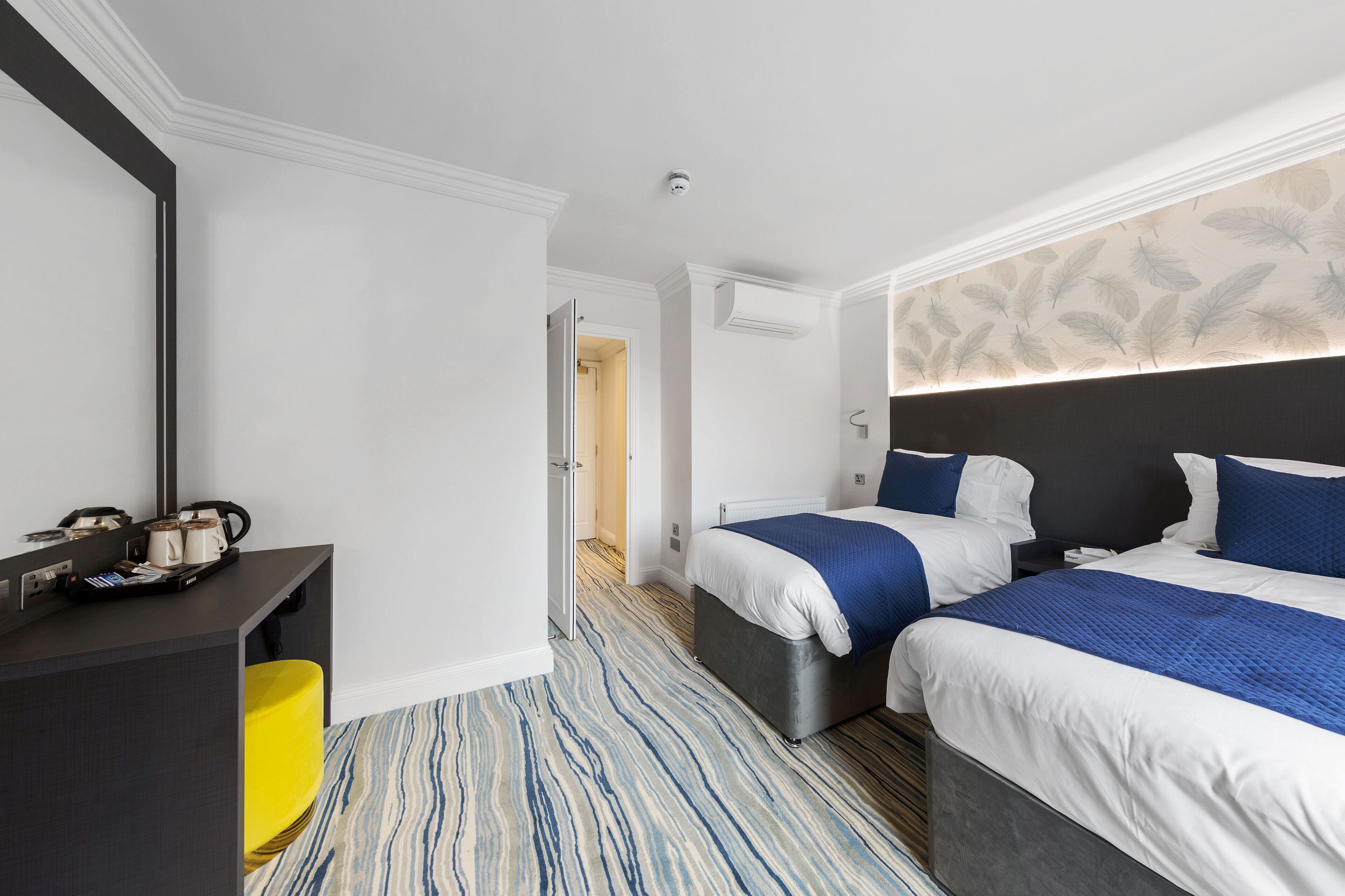 A hotel room with two beds, a desk, a chair, a trash bin, and a door leading to another room.