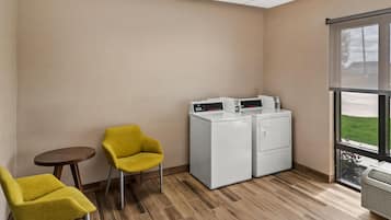 A laundry room with two washing machines, a small table, two yellow chairs, and a large window.