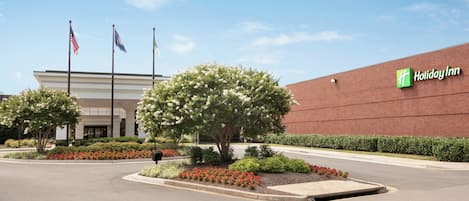 A Holiday Inn hotel with a brick facade, a landscaped entrance with flowering shrubs, and three flags.