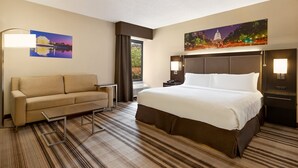 A hotel room with a bed, a sofa, a nightstand, a lamp, and a painting on the wall. - Holiday Inn Washington-Dulles International Airport by IHG (Sterling)