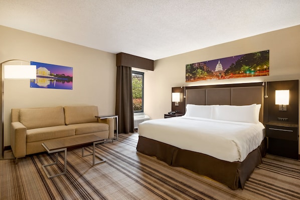 Una habitación de hotel con cama, sofá, mesita de noche, lámpara y un cuadro en la pared.