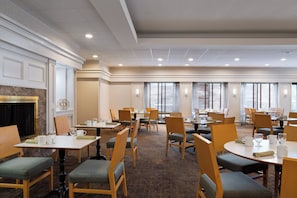 A hotel dining area with multiple tables and chairs, a fireplace, and large windows with blinds.
