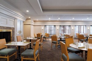 Un comedor de hotel con varias mesas y sillas, una chimenea y grandes ventanales con persianas.