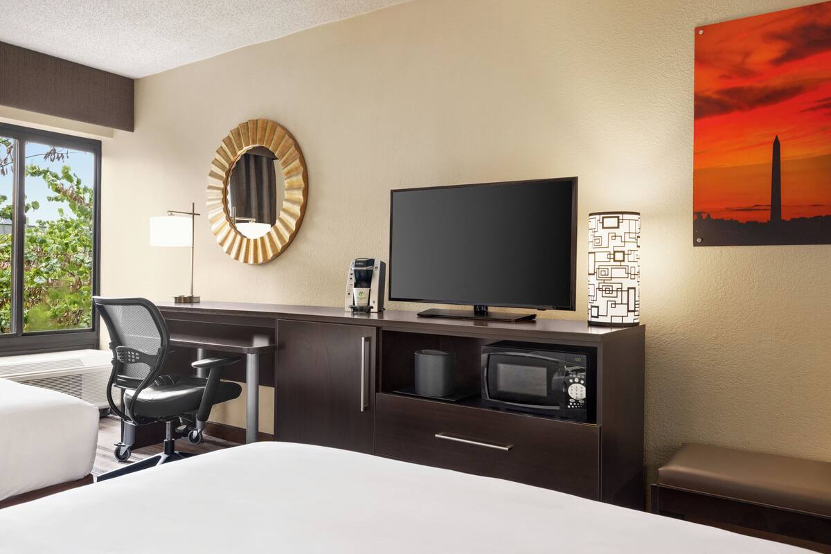 A hotel room with a bed, a desk with a computer, a TV, a coffee maker, a microwave, and a lamp.
