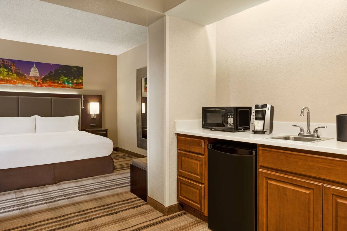 A hotel room with a bed, a kitchenette with a microwave, coffee maker, and sink, and a painting of a cityscape on the wall.