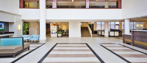 Un hall espacioso con zona central abierta, arreglos de estar, mostrador de recepción y una escalera que sube a un nivel superior.
