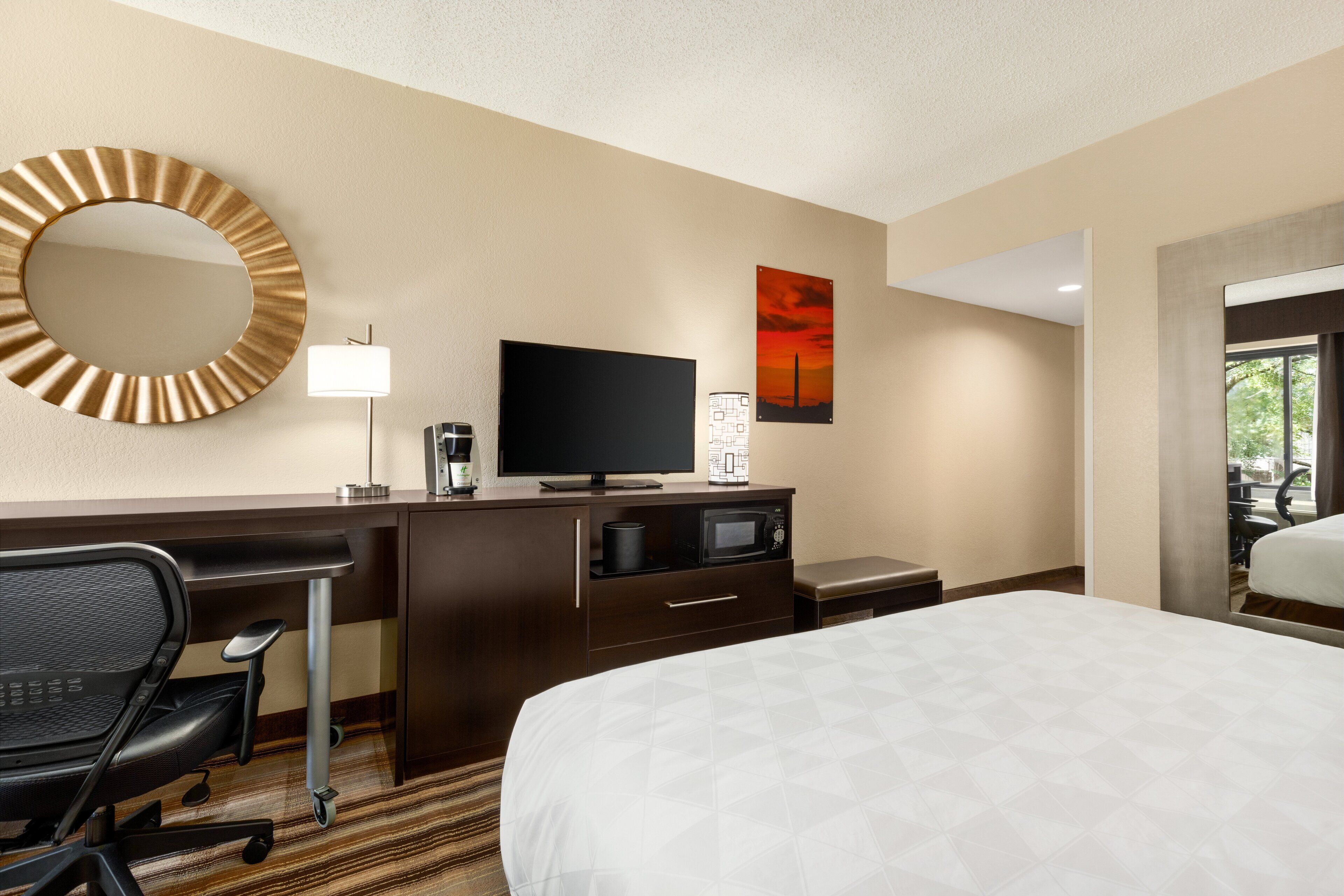 A hotel room with a bed, a desk with a chair, a television, a microwave, a coffee maker, a lamp, and a large mirror.