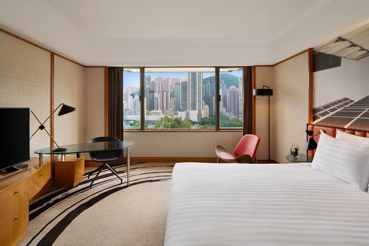 A hotel room with a bed, a desk with a chair, a TV, and a view of a cityscape through large windows.