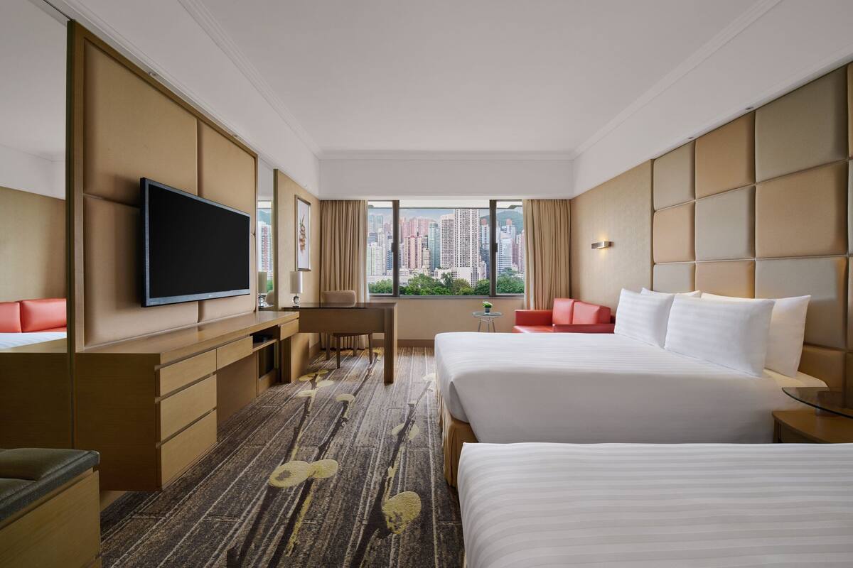 A hotel room with two beds, a desk, a television, and a view of tall buildings.
