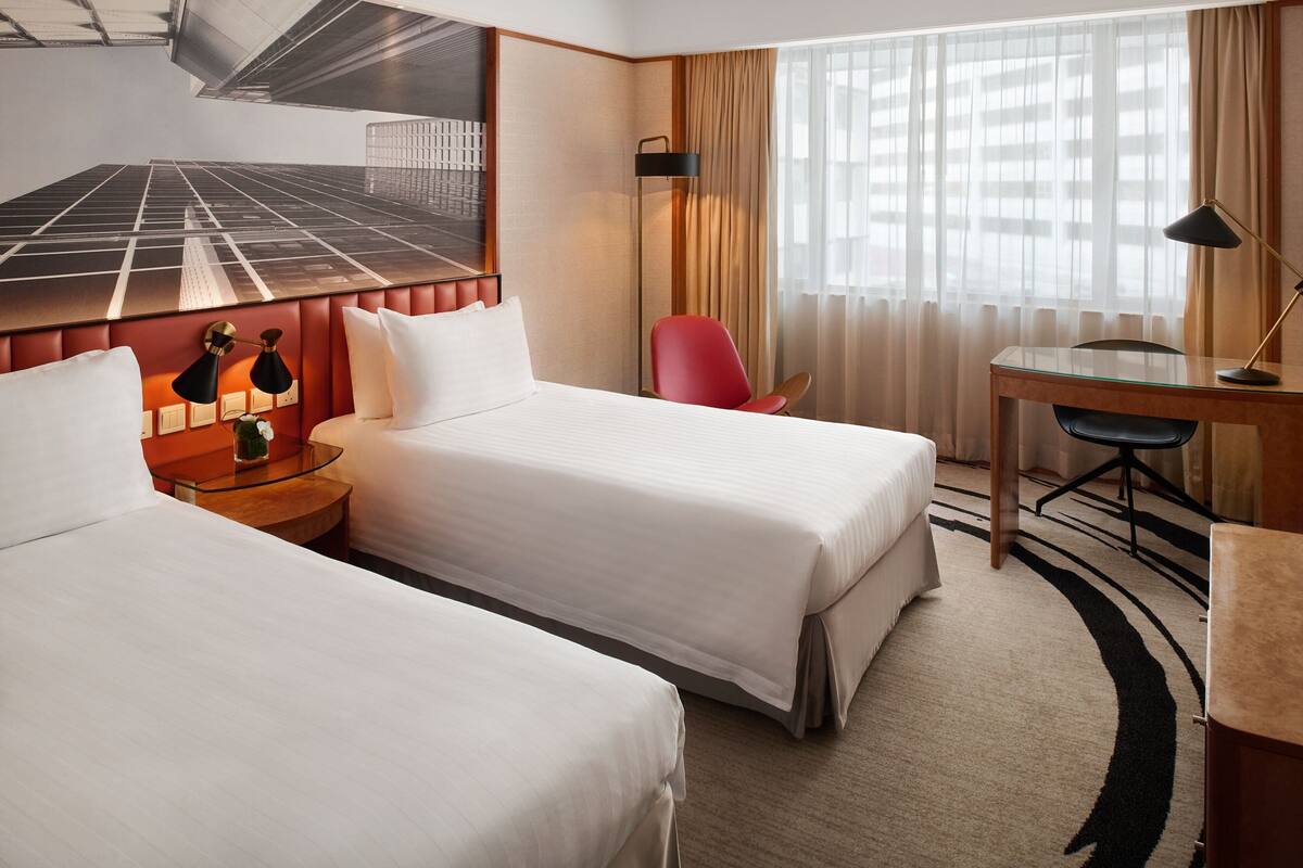 A hotel room with two beds, a desk, a chair, a lamp, and a large window with curtains.