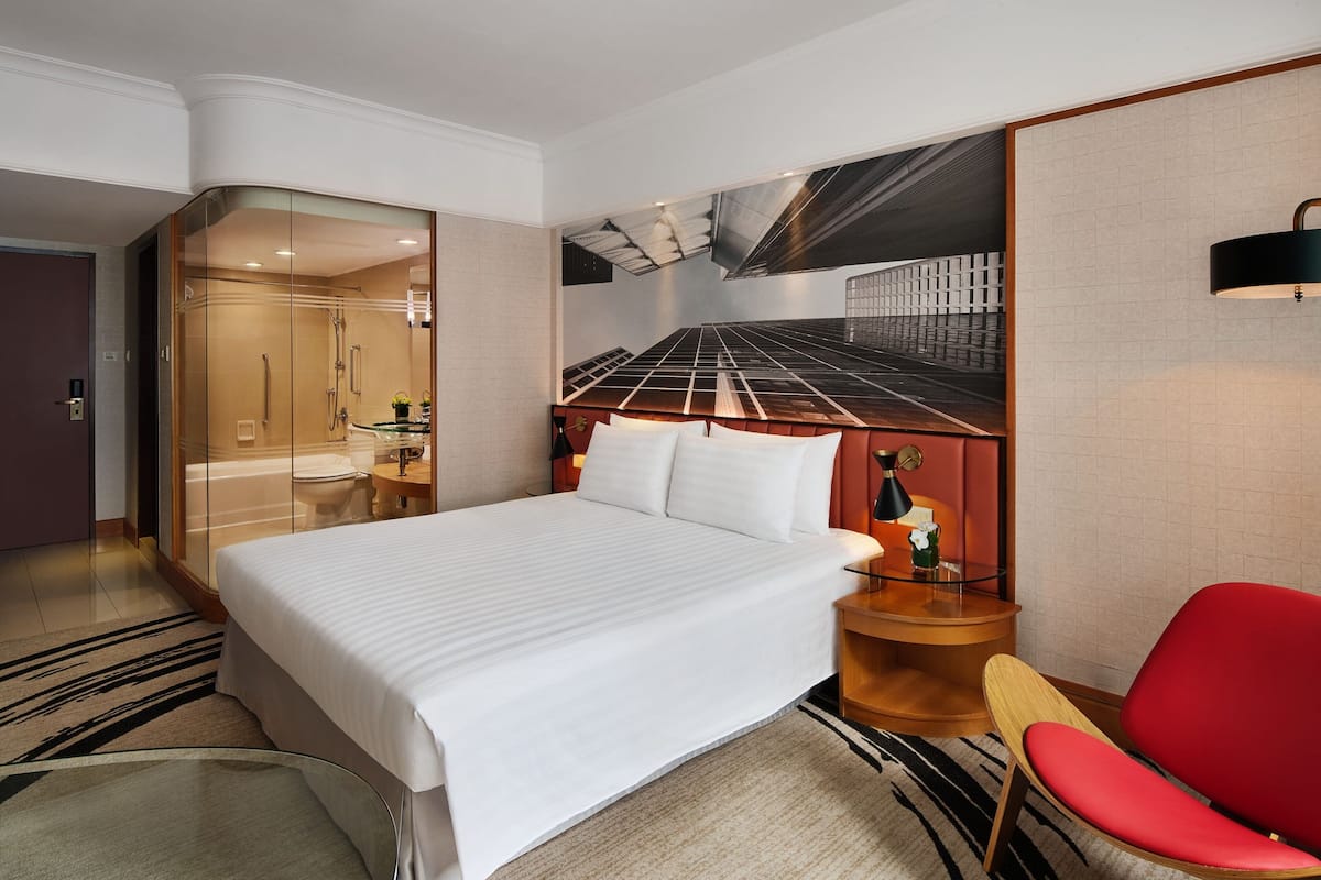 A modern hotel room with a large bed, a glass-enclosed shower, a red chair, and a cityscape view on the wall.
