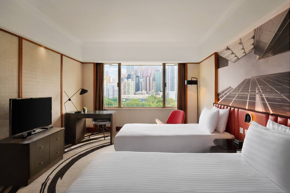 A hotel room with two beds, a desk, a TV, and a large window with a city view.