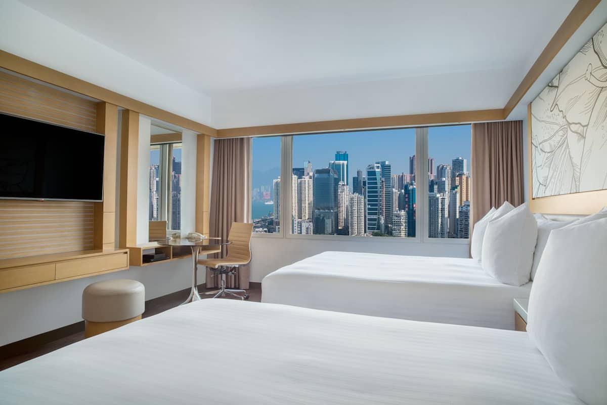 A hotel room with two beds, a desk, a chair, a TV, and a large window with a city skyline view.