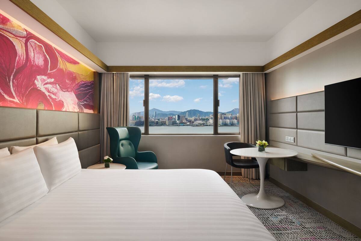 A hotel room with a large bed, a desk with a chair, a TV, and a view of a cityscape through a large window.