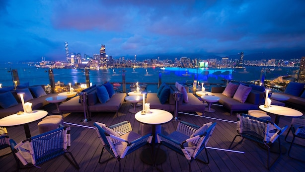 Un bar en la azotea con mobiliario exterior, incluyendo sofás y sillones, con vistas a la ciudad y al frente marítimo.
