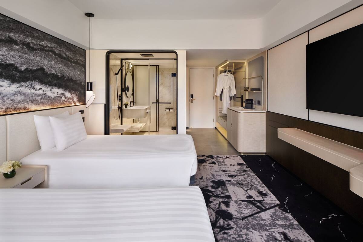 A modern hotel room with two beds, a large painting, a TV, and a bathroom with a shower and sink.