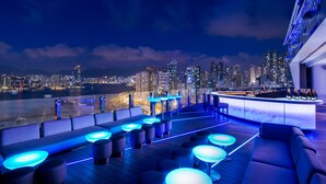 Un bar en la azotea con asientos y barra, con vistas a un horizonte urbano y una masa de agua.