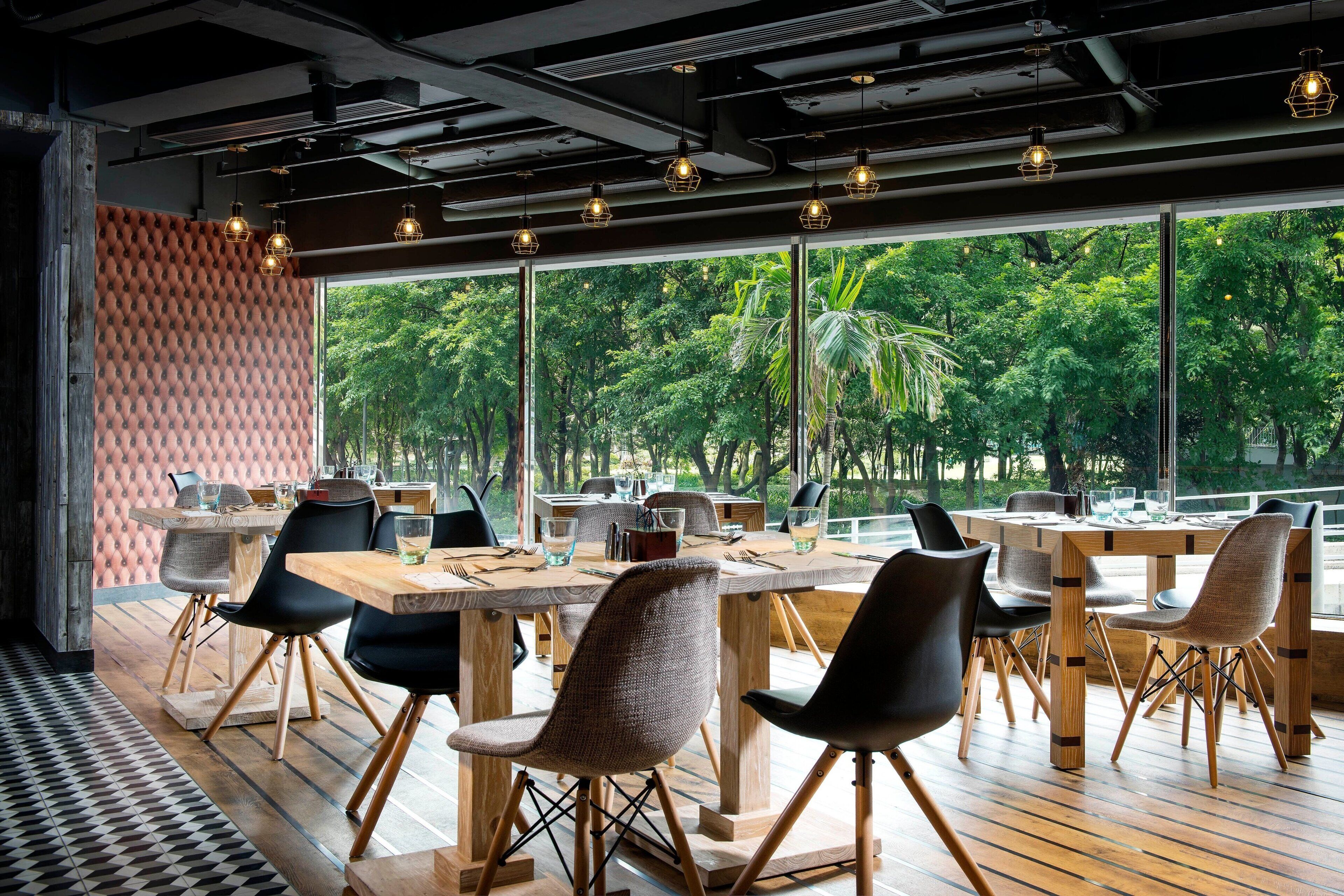 A modern dining area with wooden tables and chairs, large windows offering a view of greenery outside.