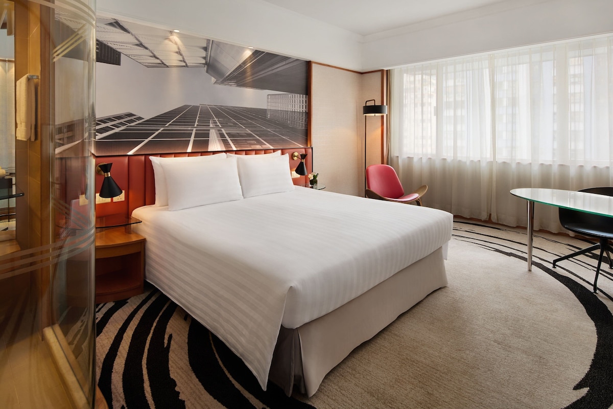 A hotel room with a large bed, a desk, a chair, and a view of the city through sheer curtains.