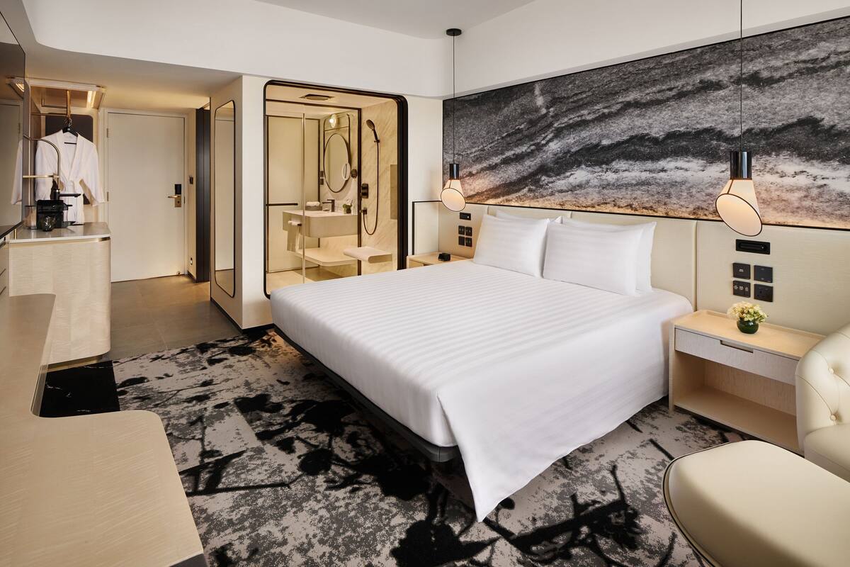 A modern hotel room with a large bed, a desk, a chair, a mirror, and a bathroom with a shower and sink.