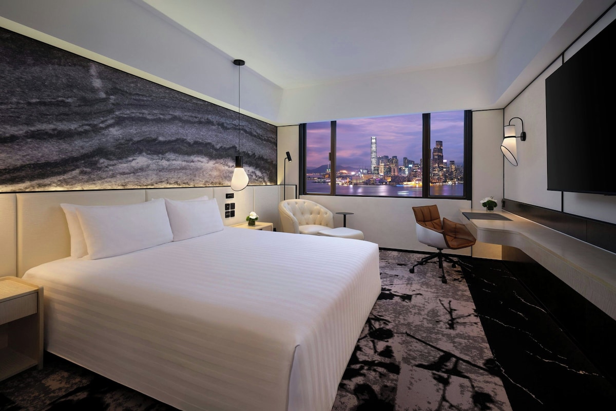 A hotel room with a bed, a desk, a chair, a TV, and a large window with a city view.