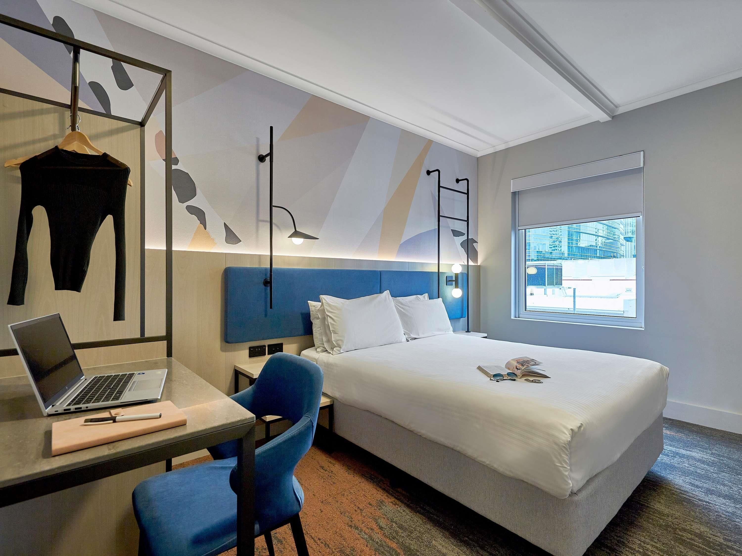 Een hotelkamer met een bed, een bureau met een laptop, een stoel en een raam met uitzicht op gebouwen.