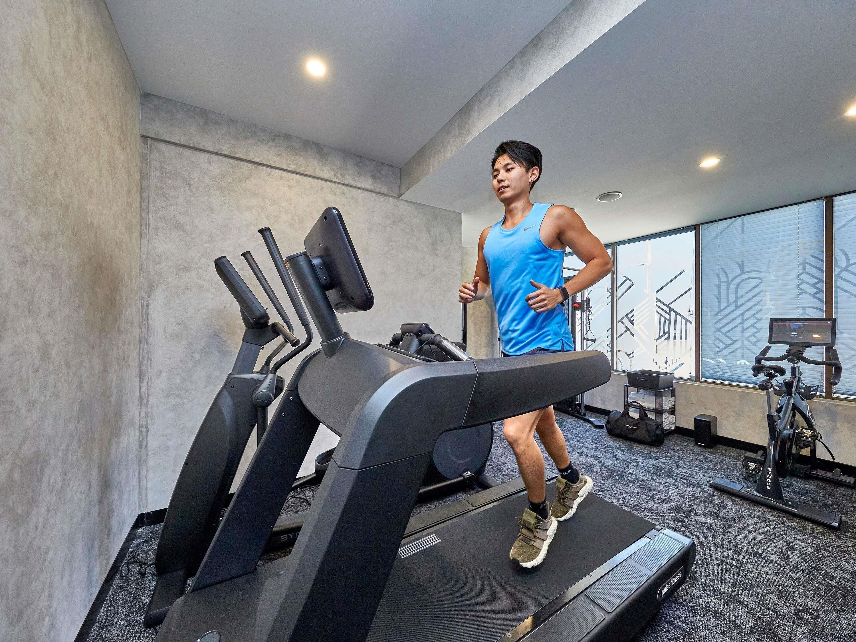 A person running on a treadmill in a gym with exercise equipment and a large window in the background.