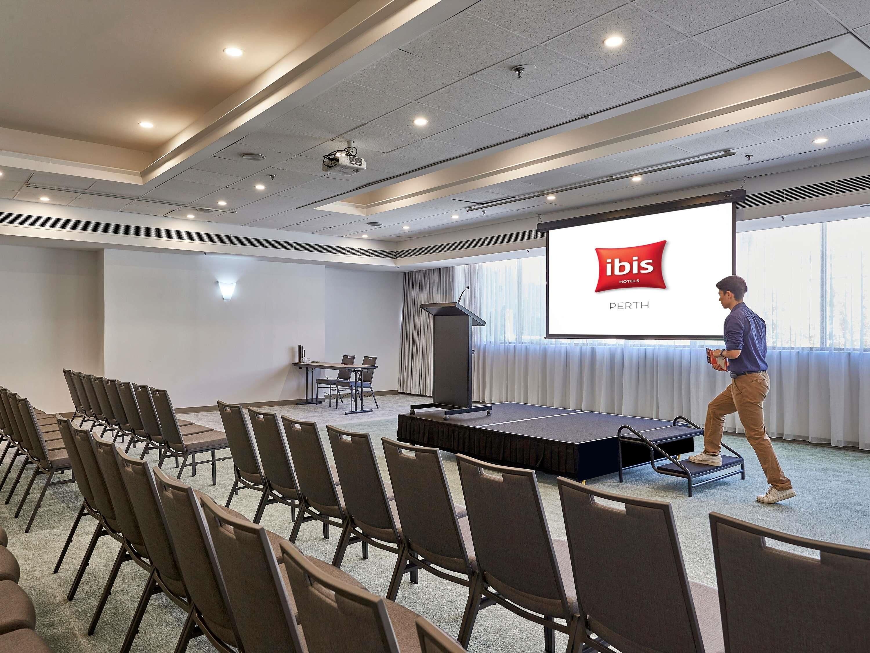 A conference room with a stage, chairs, a podium, and a large screen displaying 'ibis Hotels Perth'.
