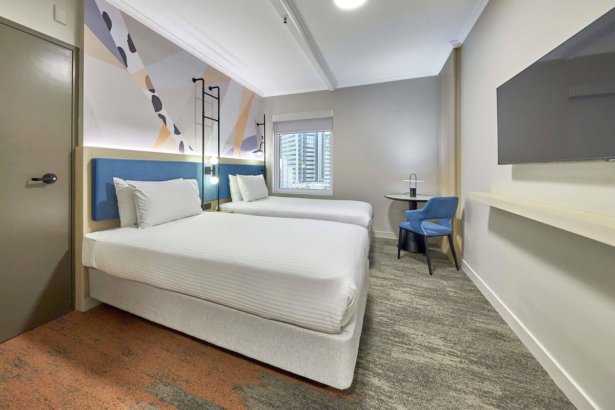 A hotel room with two beds, a desk, a chair, a TV, a window with a city view, and a ceiling light.