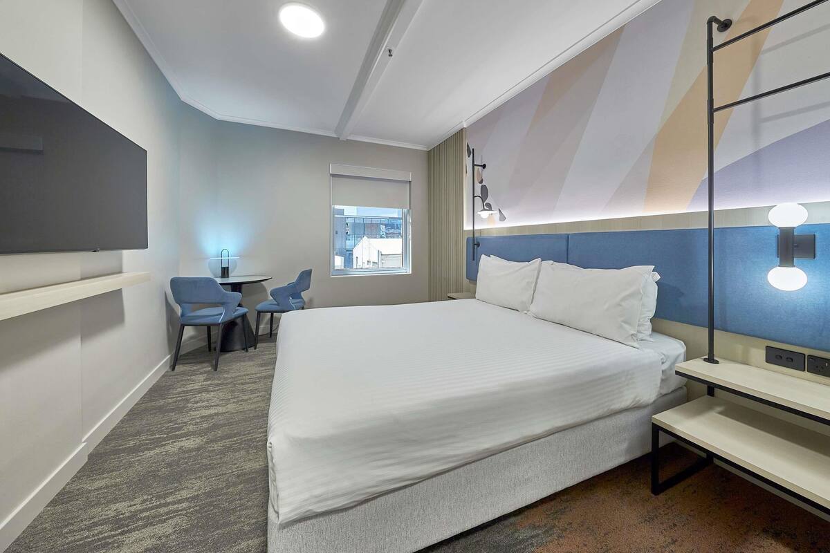 A hotel room with a bed, a desk, two chairs, a TV, and a window with a view of buildings.