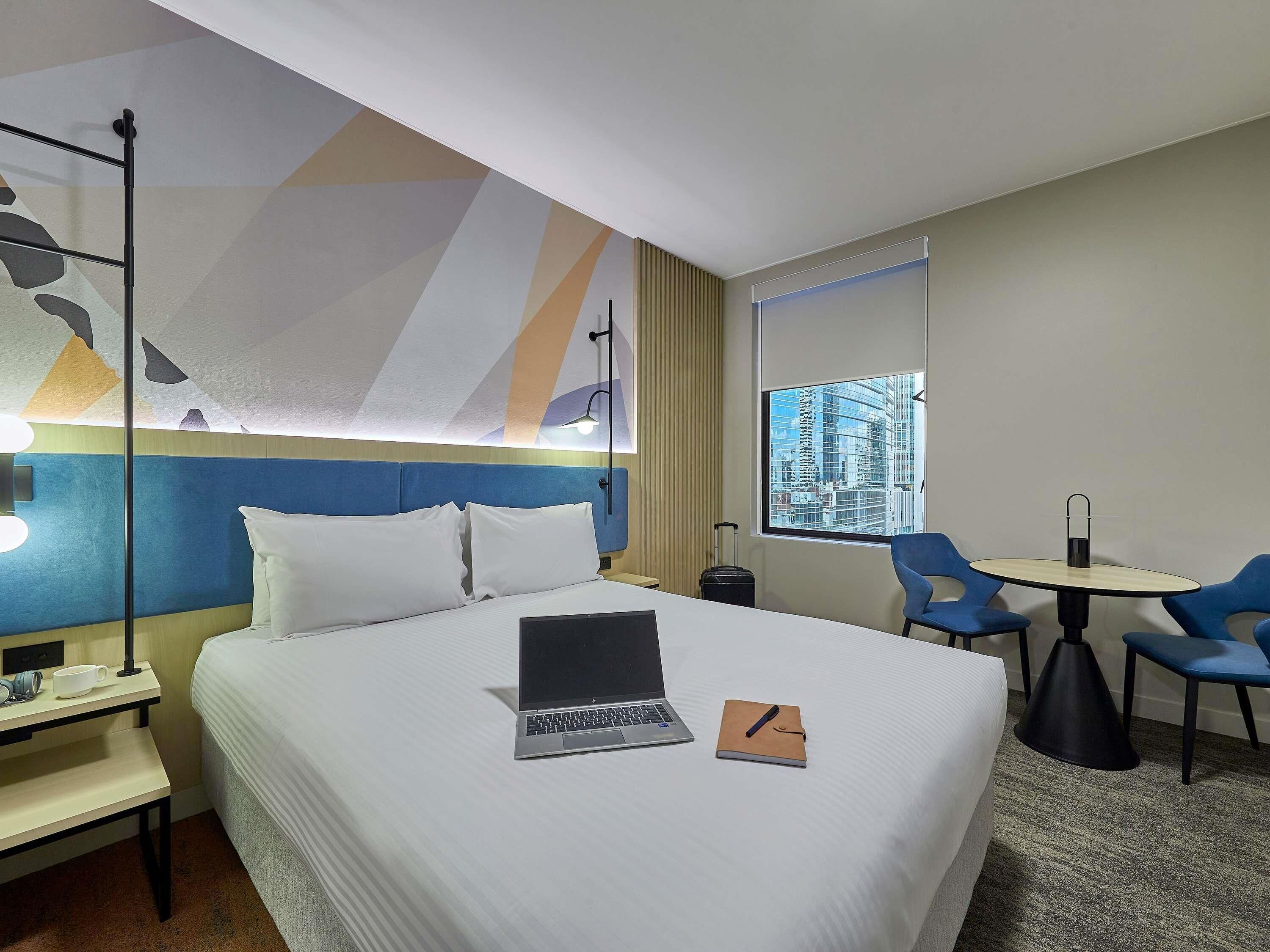 Une chambre d’hôtel avec un lit, un bureau avec un ordinateur portable, une chaise, une petite table et une fenêtre donnant sur la ville.