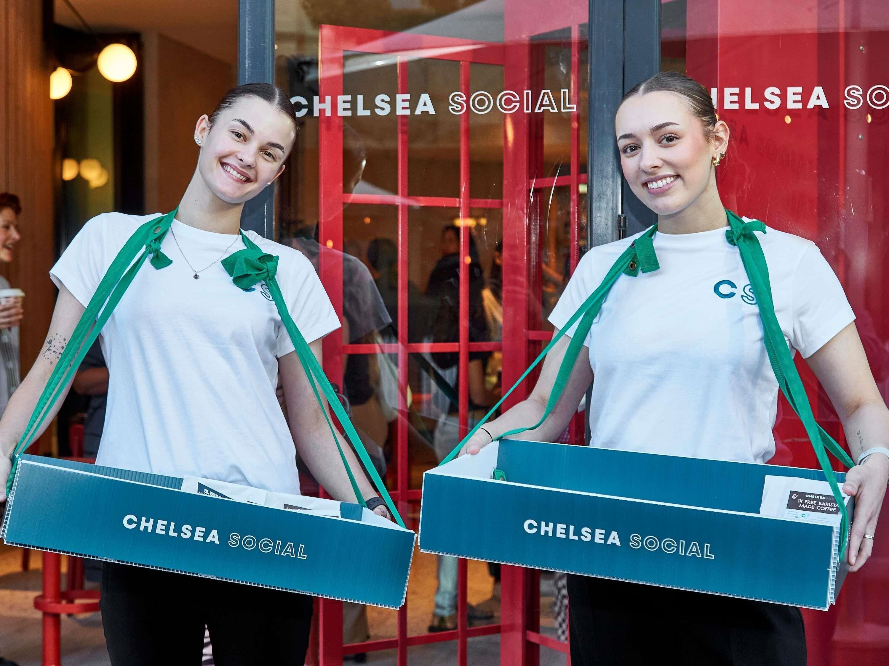 Two individuals holding Chelsea Social branded boxes outside a building with a red door and glass windows.