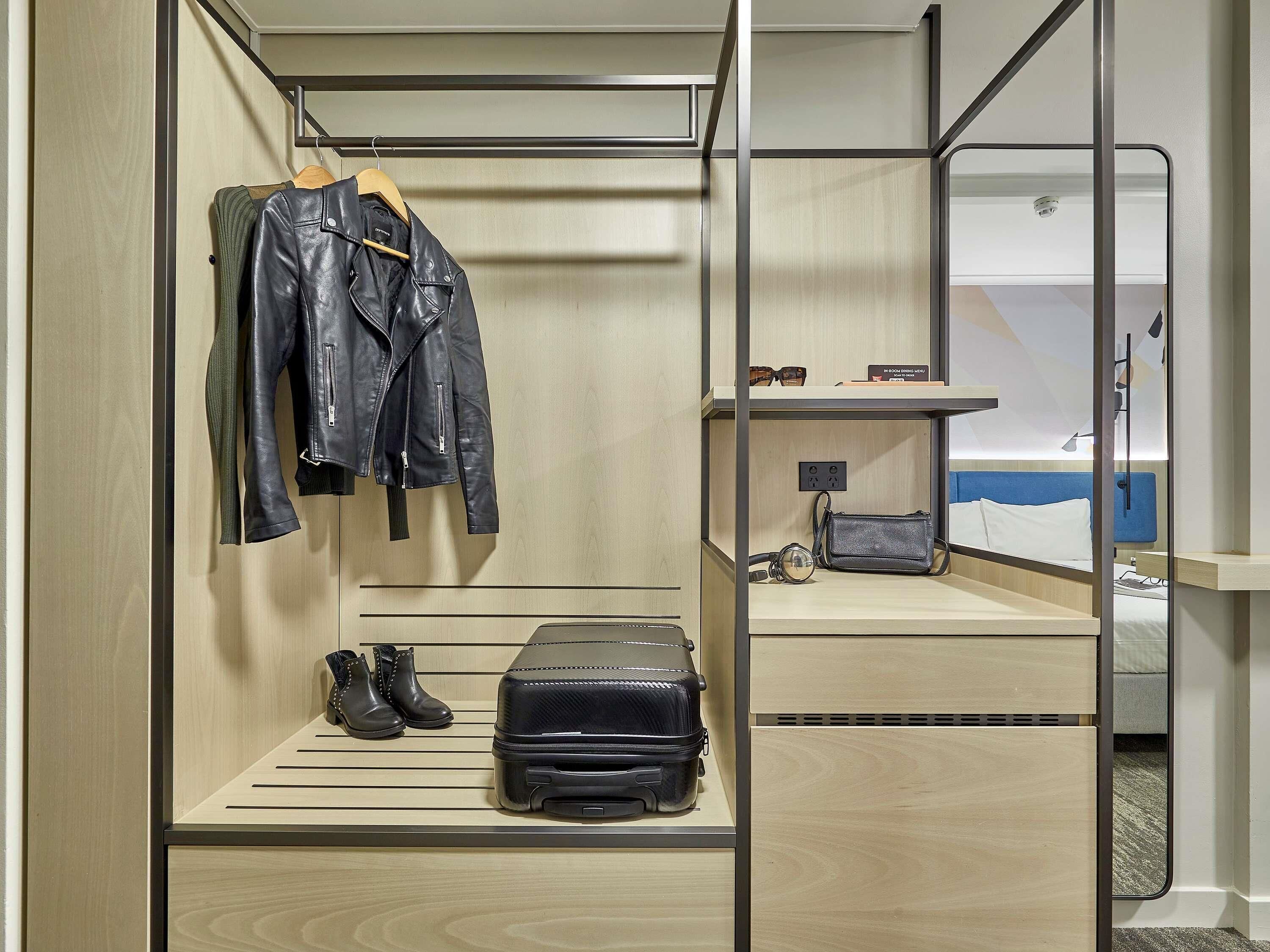 A compact hotel room with a bed, a small seating area, a mirror, and a wardrobe with hanging clothes and a suitcase.