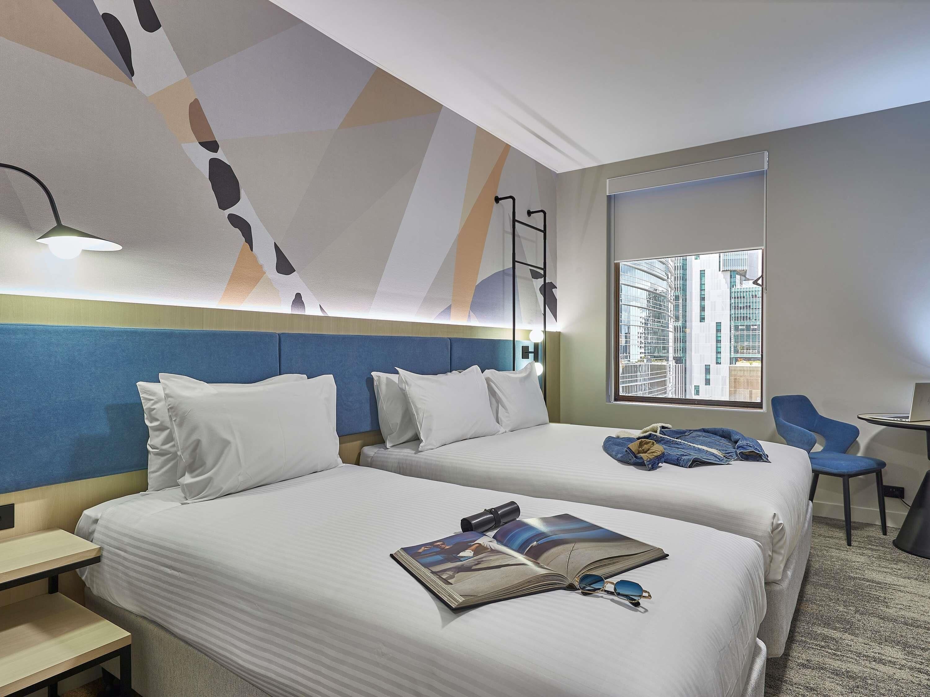 A hotel room with two beds, a desk, a chair, a lamp, and a view of a cityscape.
