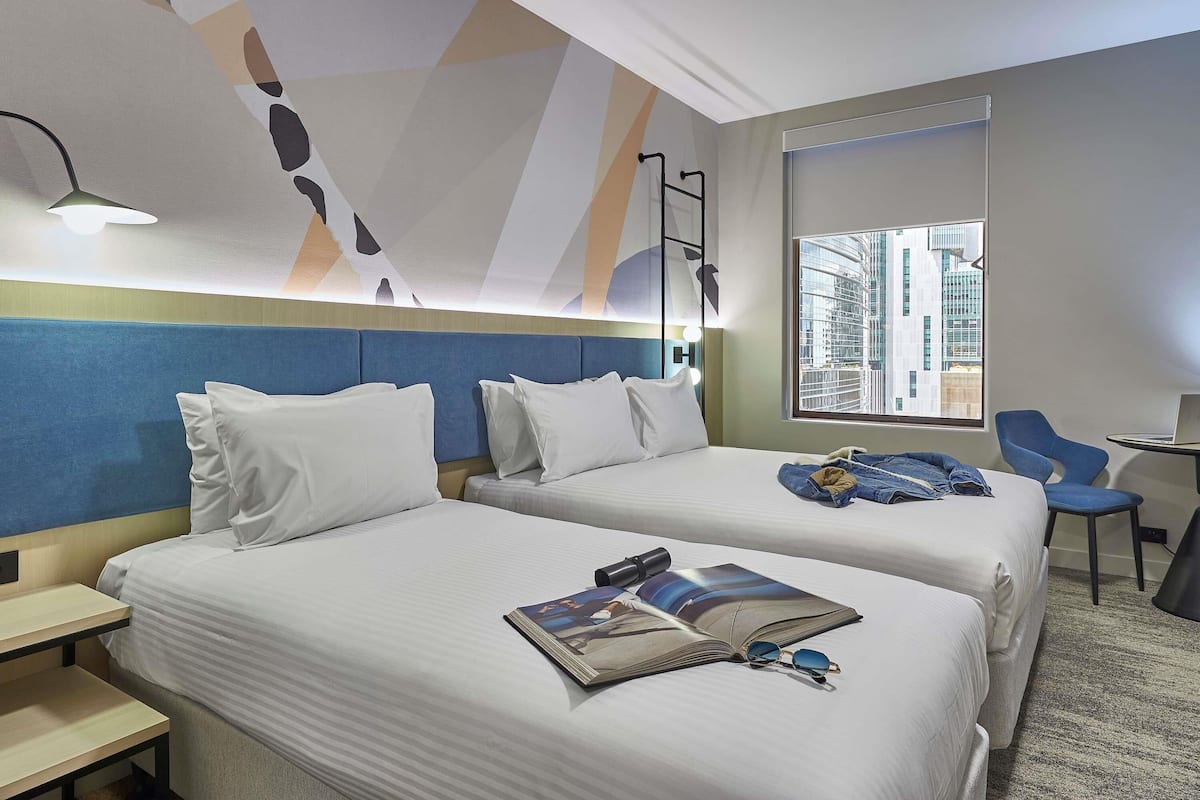 A hotel room with two beds, a desk, a chair, a lamp, and a view of a cityscape.