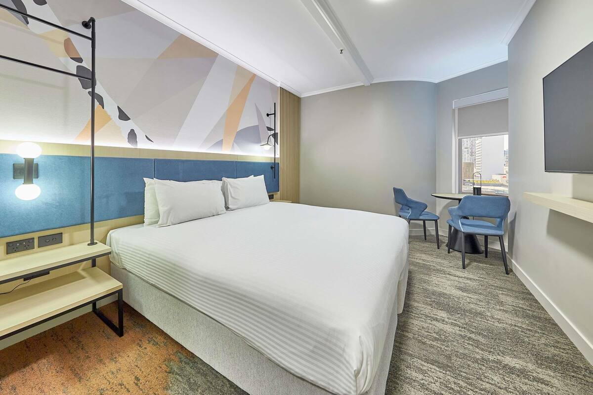 A hotel room with a bed, desk, chair, and a TV on the wall.