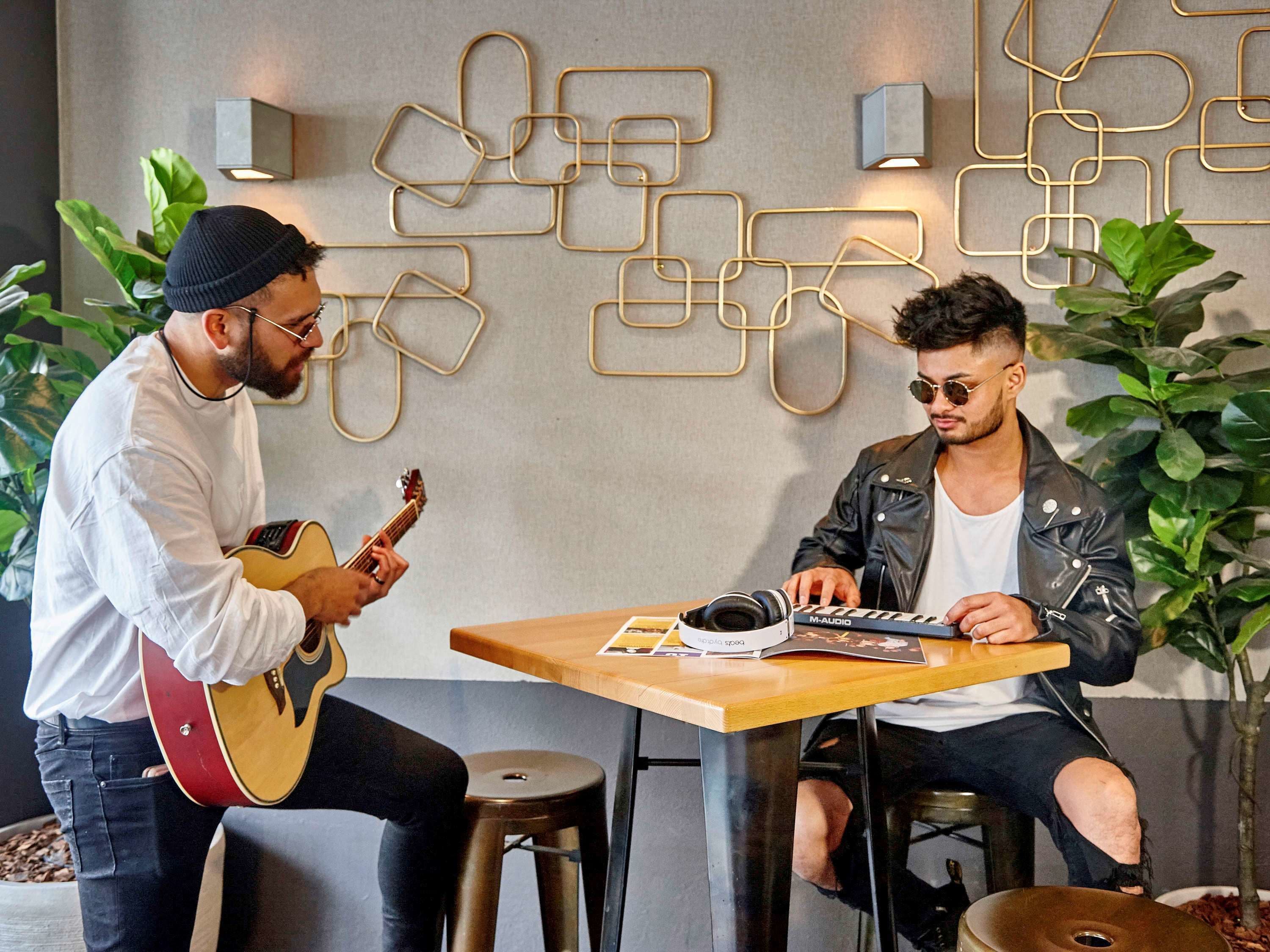Dos personas sentadas en una mesa con instrumentos musicales, rodeadas de plantas y decoración geométrica en la pared.