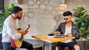 Dos personas sentadas en una mesa con instrumentos musicales, rodeadas de plantas y decoración geométrica en la pared.