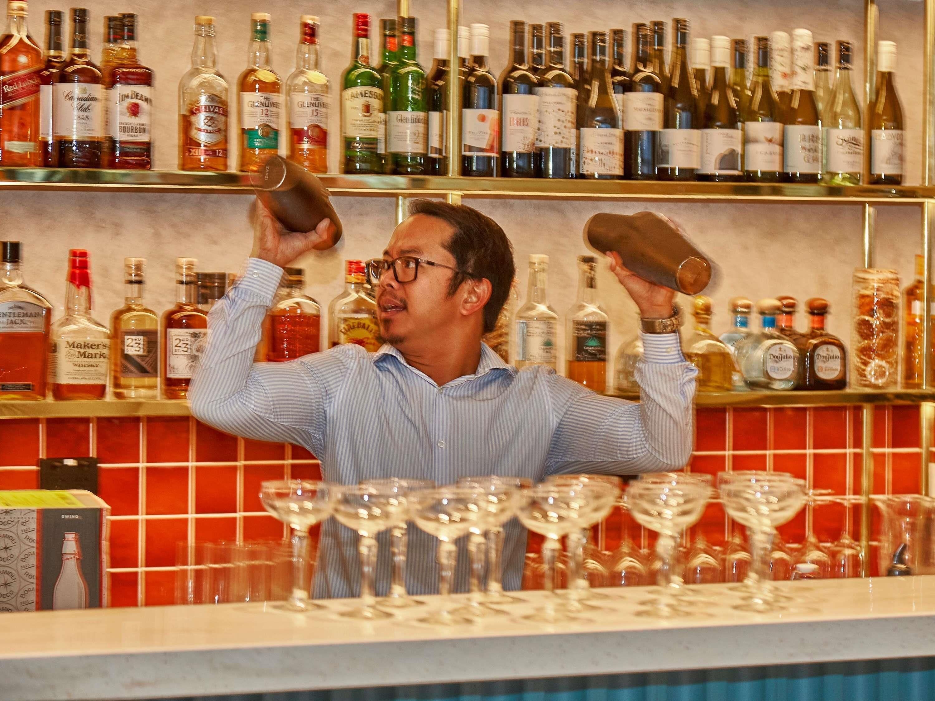A bartender holding a bottle and a shaker behind a bar with various liquor bottles and glasses.